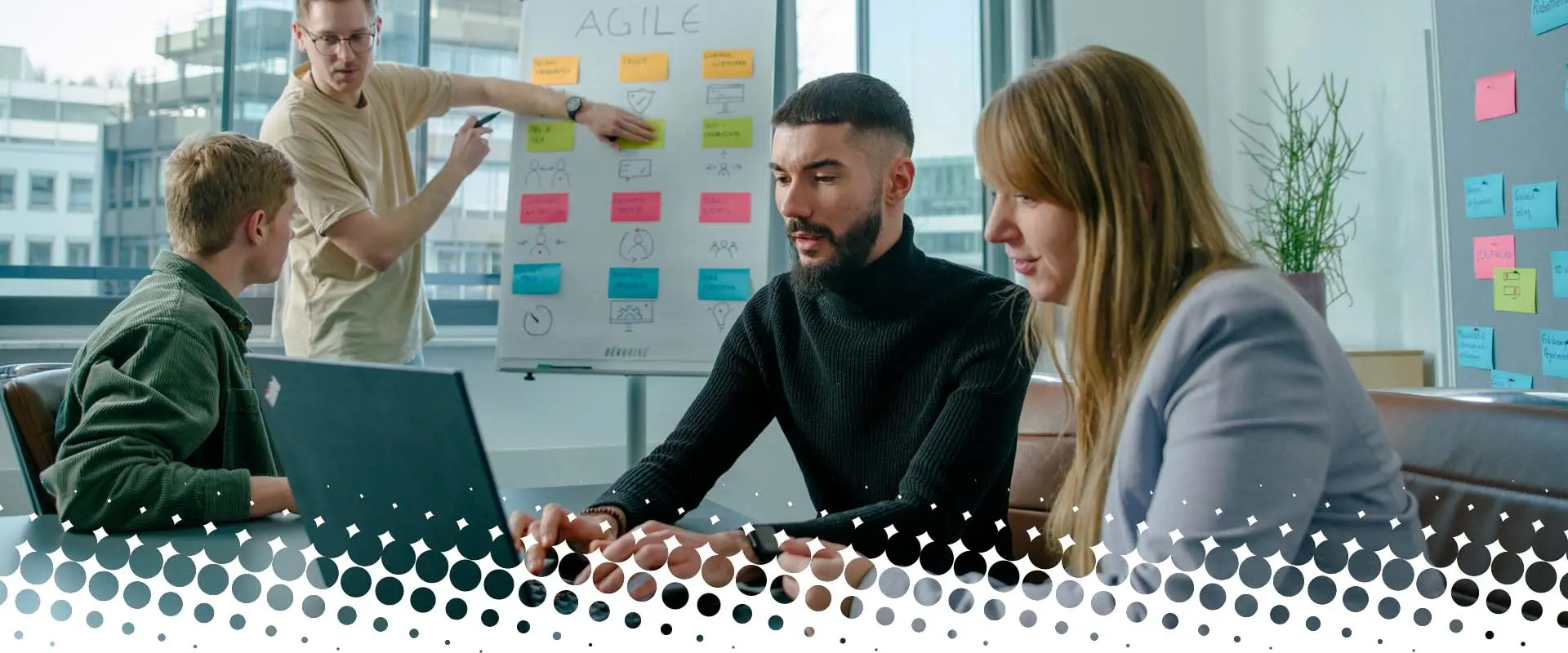 Teammeeting in einem modernen Büro: Vier Personen arbeiten zusammen, während eine Person am Whiteboard agile Methoden erklärt und andere am Laptop mitarbeiten.