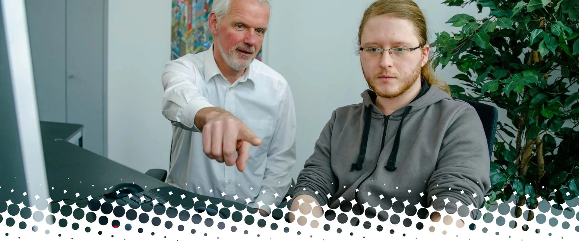 Zwei Männer sitzen in einem Büro vor einem Computer. Der ältere Mann zeigt auf den Bildschirm, der jüngere schaut geradeaus. Im Hintergrund steht eine grüne Pflanze.