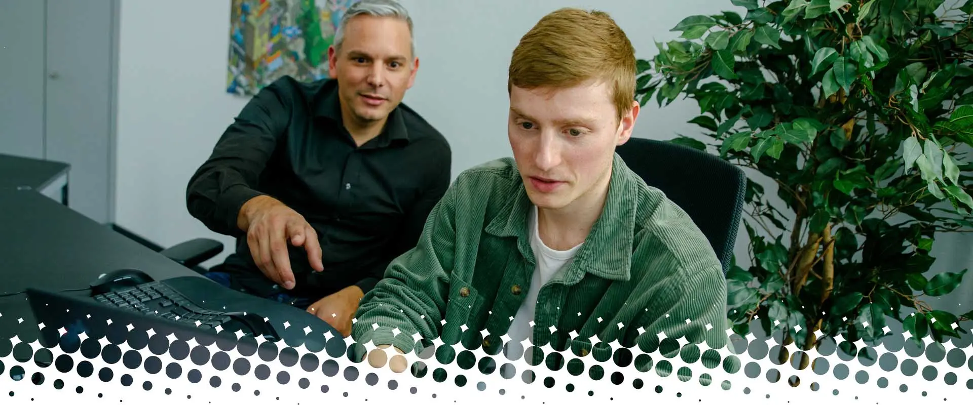 Two men are sitting in an office in front of a computer. The older man is pointing at the screen, while the younger man is looking at it with concentration. A green plant is visible in the background.