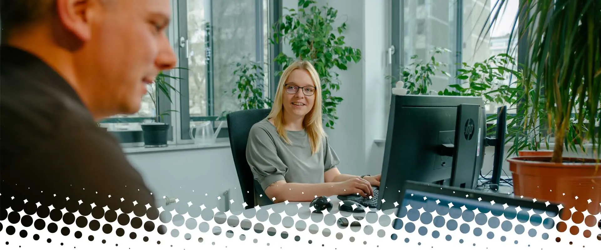 Woman sitting at a computer in an office, looking at a colleague, plants in the background.