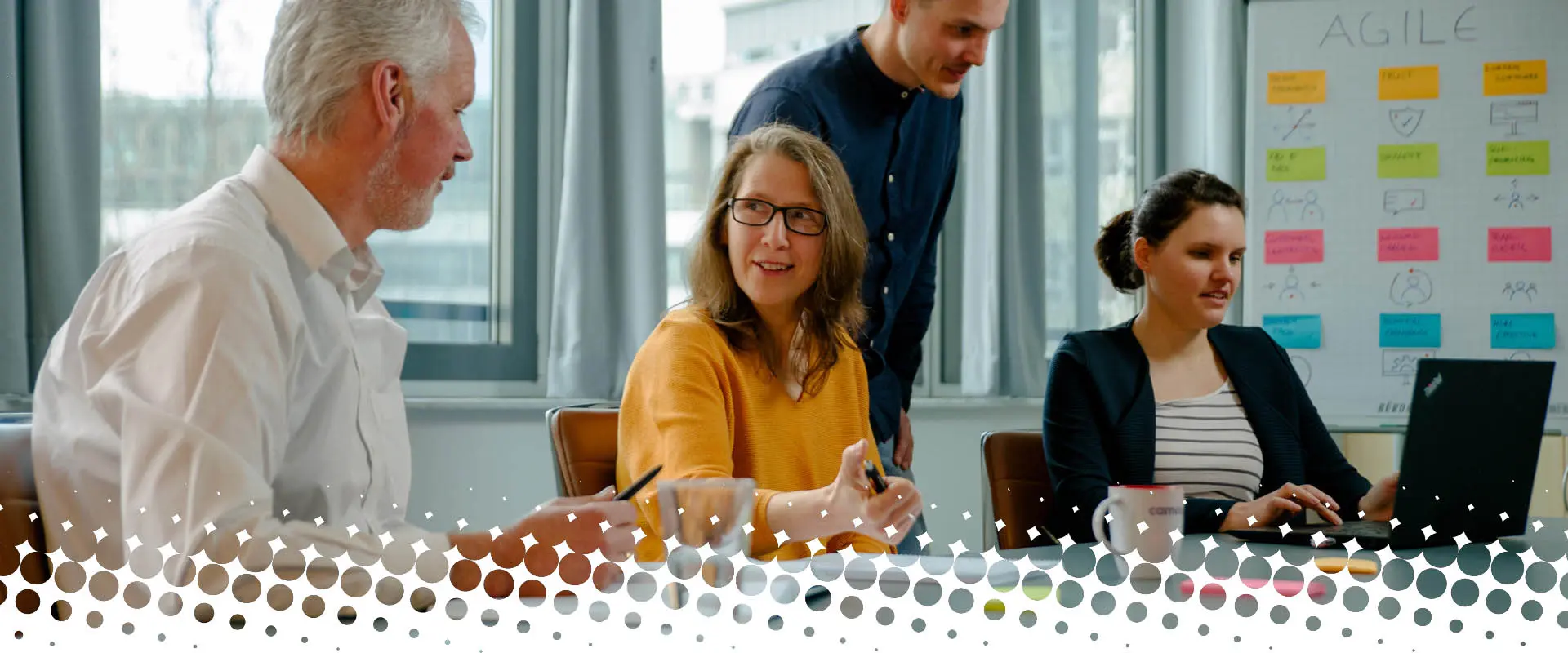 Teammeeting in einem modernen Büro: Vier camos Kolleginnen und Kollegen diskutieren und arbeiten gemeinsam an einem Tisch, im Hintergrund ist ein Whiteboard mit Agile-Methoden sichtbar.
