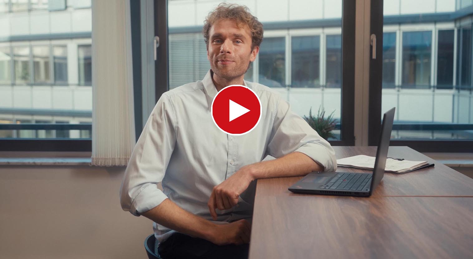 Employee sitting at a desk with a laptop in an office; a play button indicates that a video can be started.